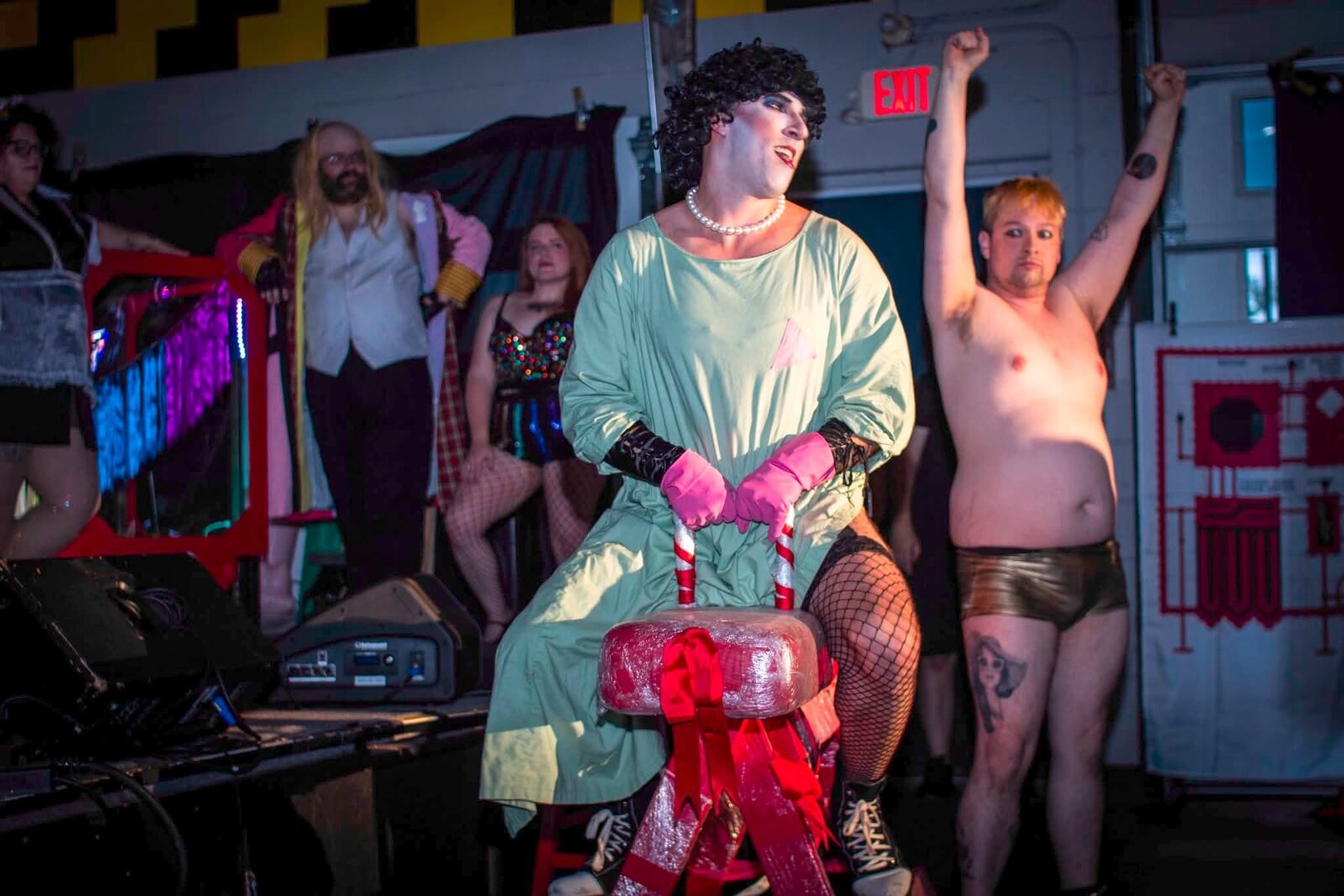 Frank-N-Furter in a green surgical gown seated on a gift-wrapped prop, cast behind