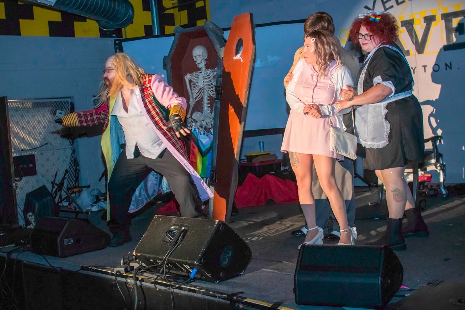 Riff Raff in a rainbow coat beside a coffin, with Janet and Magenta at Yellow Cab Tavern